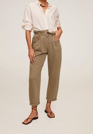 Persoon draagt beige hoge taille broek met knopen, omgeslagen zomen, een losse crèmekleurige blouse met opgerolde mouwen, bruine sandalen met bandjes en minimale sieraden.