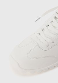 Weiße Sneaker mit glattem Synthetik-Obermaterial, kontrastierenden Nähten, runder Zehenpartie und strukturiertem Gummi-Sohle. Verfügt über flache Schnürsenkel und eine minimale Markenkennzeichnung.