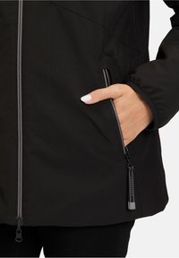 Veste noire avec fermeture à zip et poche latérale. Présente des poignets élastiques, une texture lisse et des coutures grises discrètes pour une touche de détail supplémentaire.