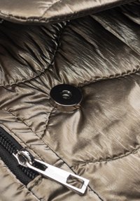 Metallisch grauer, gesteppter Stoff mit rundem Magnetknopf und teilweise sichtbarem schwarzem Reißverschluss mit silbernem Zippergriff.