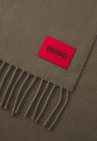Oliven grøn tørklæde med et rødt mærke med "HUGO" i sort. Blød tekstur med frynset kanter og et vævet mønster.