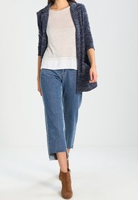 Námořnický pletený cardigan přes světlejší top, spárovaný s modrými zkrácenými džíny a hnědými kotníkovými botami. Má uvolněný střih a kombinaci různých textur.