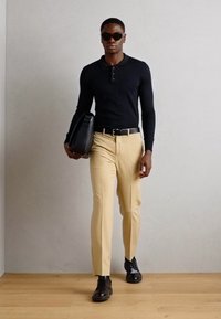 Juodas ilgomis rankovėmis polo marškinėliai, smėliniai aptempti kelnės, juodi batai, pilka kuprinė ir saulės akiniai, prieš lengvai pilką sieną ir medinį grindų paviršių.
