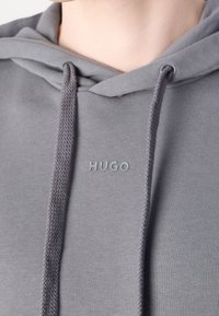 Gros plan d'un sweat à capuche gris clair avec cordons et "HUGO" imprimé en petits caractères en relief sur la poitrine.