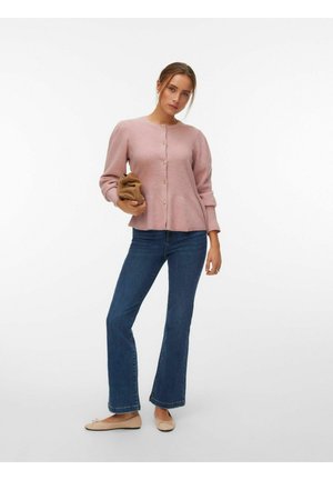 Femme debout portant un cardigan rose clair boutonné, un jean bleu foncé, des ballerines beiges, et tenant une pochette marron pliée sur fond blanc.