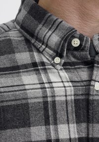 Flanellskjorte med knapper i svart-hvitt rutete mønster og teksturert stoff, med spredd krage og hvite knapper. Lukket kragedetalj synlig.