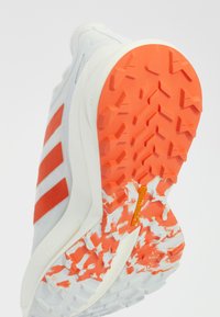 White athletic shoe with red stripes, textured surface, and a rubber outsole featuring an orange and white pattern. Contoured design for support.