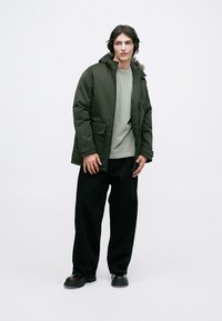 Khaki pufferjakke med hætte, forlommer, grå sweatshirt og sorte brede bukser, parret med sorte sko. Enkel, funktionel design.