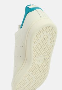 Sneaker krémové barvy s tyrkysově semišovým límcem, designem bez tkaniček, perforovanými detaily a texturovanou gumovou podrážkou s vlnkovým vzorem.