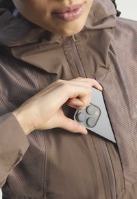 Main tenant un smartphone, le plaçant dans la poche poitrine d'une veste marron zippée.