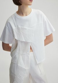 White short-sleeve t-shirt with layered fabric panels featuring varying textures. Clean lines and a loose fit create a contemporary design.