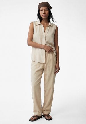 Femme portant une chemise sans manches beige à boutons, un pantalon ample assorti, un foulard marron sur la tête et des sandales noires, debout devant un fond blanc.