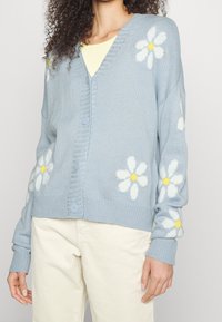 Žena oblečená v svetle modré cardiganové sako s bílými a žlutými vzory sedmikrásek, doplněné krémovými kordovým kalhotami a bledě žlutým topem.