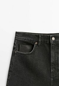 Jeans en denim noir avec une poche avant contenant un petit portefeuille en cuir noir, bouton et passant de ceinture visibles sur la taille.