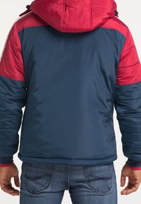 Veste matelassée zippée marine et rouge avec capuche, présentant des épaules matelassées et une texture lisse. Jean en denim bleu visible en dessous de la veste.