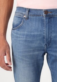 Jeans de mezclilla azul con un lavado ligeramente deslavado, diseño de cinco bolsillos, costuras en contraste y un botón de metal en la cintura.