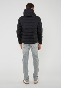 Hombre vestido con una chaqueta de plumas negra con capucha, jeans gris claro y zapatillas blancas, de pie de espaldas contra un fondo liso.