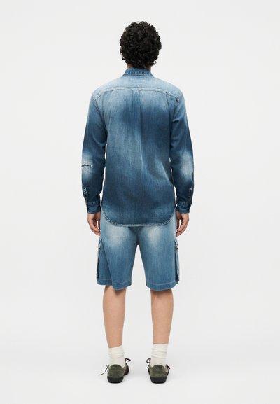 Άντρας με σγουρά μαλλιά που στέκεται πλάτη, φοράει ξεθωριασμένο μπλε τζιν πουκάμισο, ανδρικά τζιν σορτς μέχρι το γόνατο, λευκές κάλτσες και σκούρα παπούτσια.