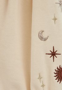 Tessuto a maglia beige con ricami di sole, luna, stelle e linee in filo marrone e oro. Superficie texturizzata con un aspetto morbido e accogliente.