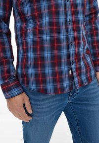 Rot, blau und marine-karierter Hemd mit langen Ärmeln und Knopfverschluss. Verfügt über einen abgerundeten Saum und schwarze Knöpfe. Getragen mit blauen Jeans.