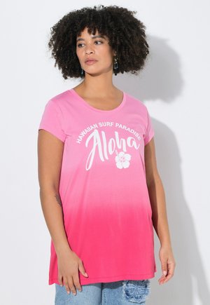 Femme aux cheveux bouclés portant un T-shirt ombré rose avec le texte "Hawaiian Surf Paradise Aloha" et un motif d'hibiscus, associée à un jean bleu.