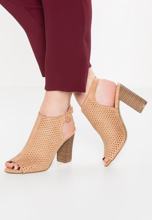 Beige perforerede peep-toe høje hælsandaler med ankelremme båret med bordeaux cropped bukser på hvid baggrund.