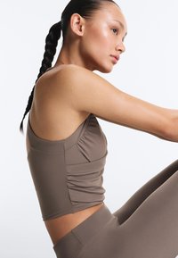 Mladá žena s vrkočmi, oblečená v taupe bezrukávovom topu a legínach, sedí so zdvihnutými kolenami na jednoduchom bielom pozadí.