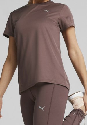 Femme étirant la jambe, portant un haut de sport et un legging marron assortis de la marque Puma, tenant une basket beige derrière son dos.