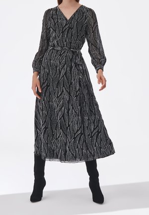 Weibliches Model trägt schwarzes durchsichtiges Midikleid mit weißen Blattmustern, langen Ärmeln, V-Ausschnitt, Taillengürtel und schwarzen Stiefeln mit hohen Absätzen.