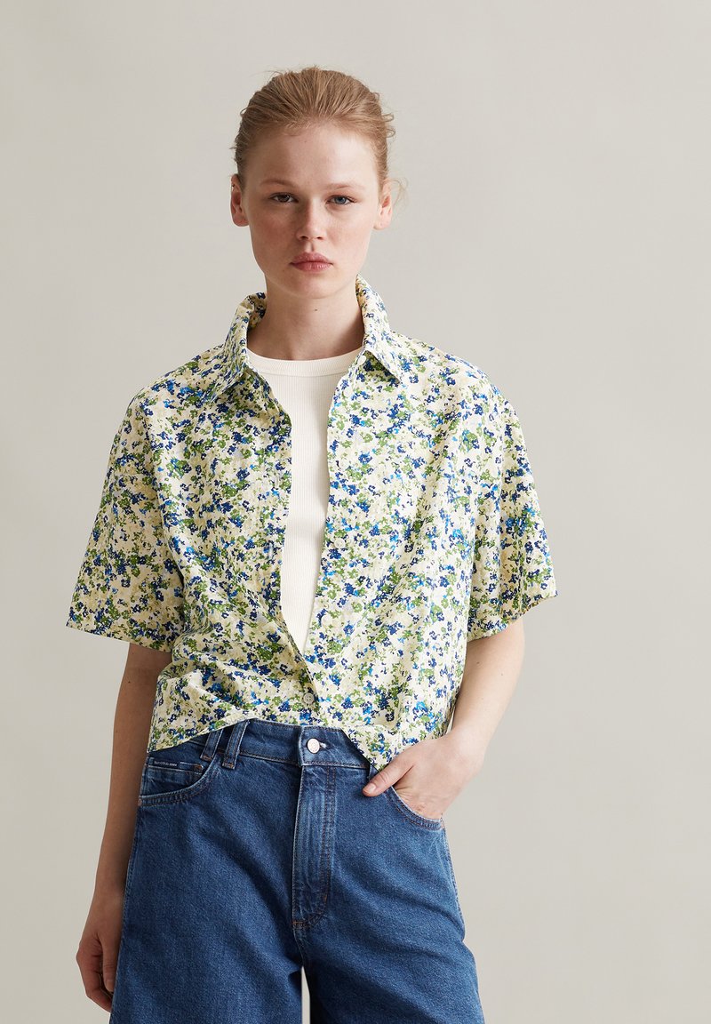 Blumen Kurzarmhemd mit Button-Down-Kragen, das grüne und blaue Blumen auf hellbeigem Hintergrund zeigt, getragen über einem weißen Oberteil.