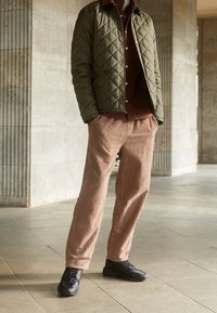 Vadderad olivgrön jacka över en brun skjorta. Ljust bruna kordflöjtbyxor med vertikala ränder och svarta sneakers. Släta texturer genomgående.