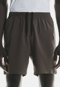 Mann in dunkelbraunen, elastischen Shorts mit schwarzen Kordeln und einem weißen Hemd, der entspannt mit den Händen an den Seiten steht.
