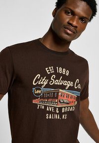 Camiseta de algodón marrón con una impresión gráfica de un edificio y el texto "City Salvage Co." Cuello redondo y mangas cortas.