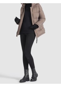 Abrigo tipo puffer en color topo con capucha, suéter negro debajo, mallas negras y botas de tobillo negras con suelas gruesas, con cierre de cremallera.