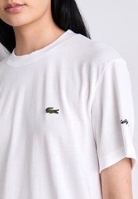 Primer plano de una persona que lleva una camiseta blanca sencilla con un pequeño logotipo verde de cocodrilo en el pecho y texto negro parcial en la manga.