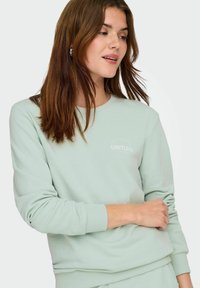 Lysegrønn sweatshirt laget av mykt stoff, med rund hals, ribbede mansjetter og en liten "LIMITLESS" logo på forsiden.