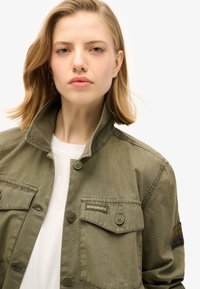 Olijfgroene jas van lichtgewicht stof, met een omgeslagen kraag, knopen aan de voorkant en twee borstzakken met logo-detail.