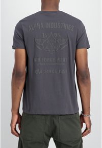 Camiseta de algodón gris con un gráfico en la parte posterior que dice "ALPHA INDUSTRIES AIR FORCE PILOT" junto con un diseño de espada alada. Mangas cortas, cuello redondo.