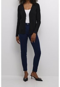 Svart långärmad cardigan över en vit linne, kombinerad med mörkblå skinny jeans och svarta klackskor med dekorationer.