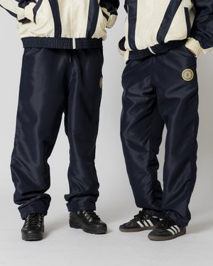 Pantaloni de trening din nailon albastru marin, cu talie elastică și mansete, prevăzuți cu un accent circular cu logo, asortați cu cizme negre și adidași.