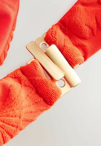 Rode stoffen band met een gestructureerd patroon en een gouden metalen sluiting met twee gaten. Het materiaal lijkt zacht en geweven.