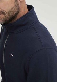 Mornarica majica sa zipom koja ima visoki ovratnik, glatku teksturu i mali trobojni logo na prsima.