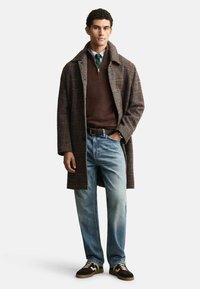 Wollmischung-Überjacke in braunem Karomuster, getragen über einem braunen Pullover, blauen Jeans und dunklen Sneakers mit einem weißen Muster. Elegante Passform.