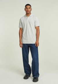 camiseta polo gris claro de manga corta, con cuello plano y un sutil logo en el lado izquierdo, combinada con unos jeans de pierna ancha azul oscuro y zapatos negros.