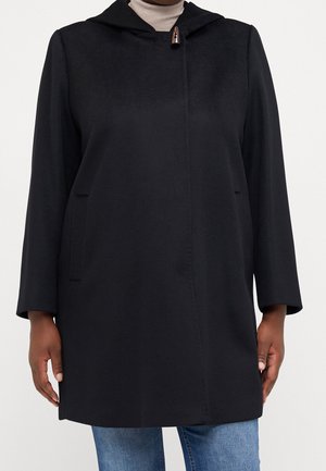 Manteau noir mi-long avec capuche et fermeture à boutons à bascule, porté sur un col roulé beige et un jean bleu, sur une personne à peau foncée.