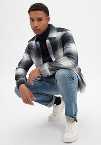 Gestreepte shirt in marineblauw, wit en lichtblauw, met knoopsluiting, gedragen over een marineblauwe coltrui, gecombineerd met lichtblauwe jeans en witte sneakers.