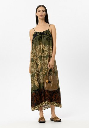 Woman with long dark hair wearing a green floral maxi dress holding a brown leather handbag and wearing brown open-toe sandals.