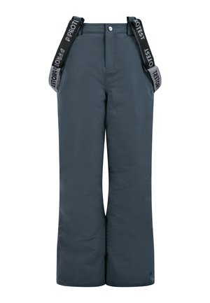Jeune personne portant un bonnet en tricot bleu, une polaire bleu clair zippée, un pantalon de neige foncé avec des bretelles et des bottes noires, debout avec une expression neutre.