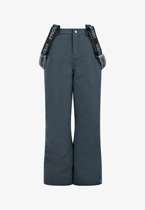 Jeune personne portant un bonnet en tricot bleu, une polaire bleu clair zippée, un pantalon de neige foncé avec des bretelles et des bottes noires, debout avec une expression neutre.