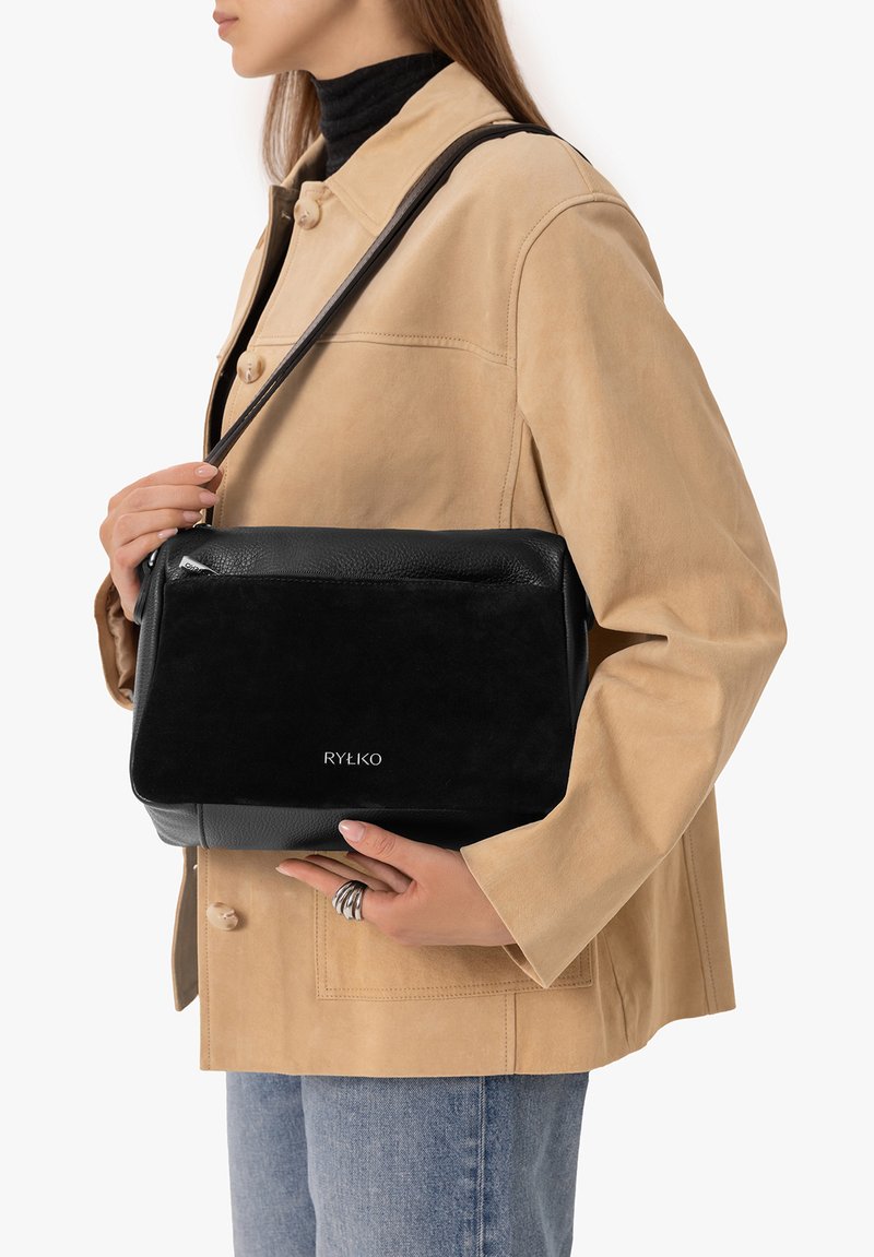 Femme en veste beige et jean bleu tenant un sac à bandoulière noir RYŁKO avec une fermeture éclair argentée et plusieurs bagues aux doigts.